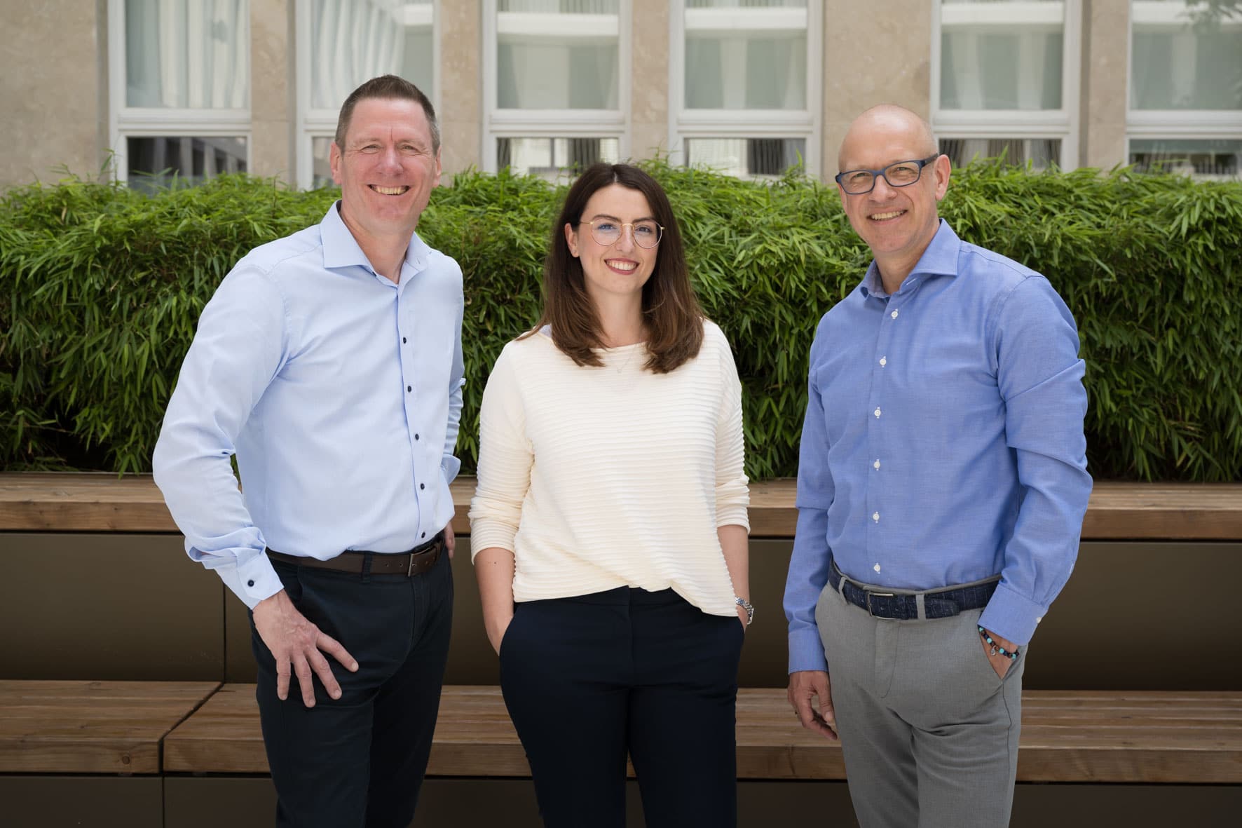 Eine Frau und zwei Männer lächeln draußen in die Kamera