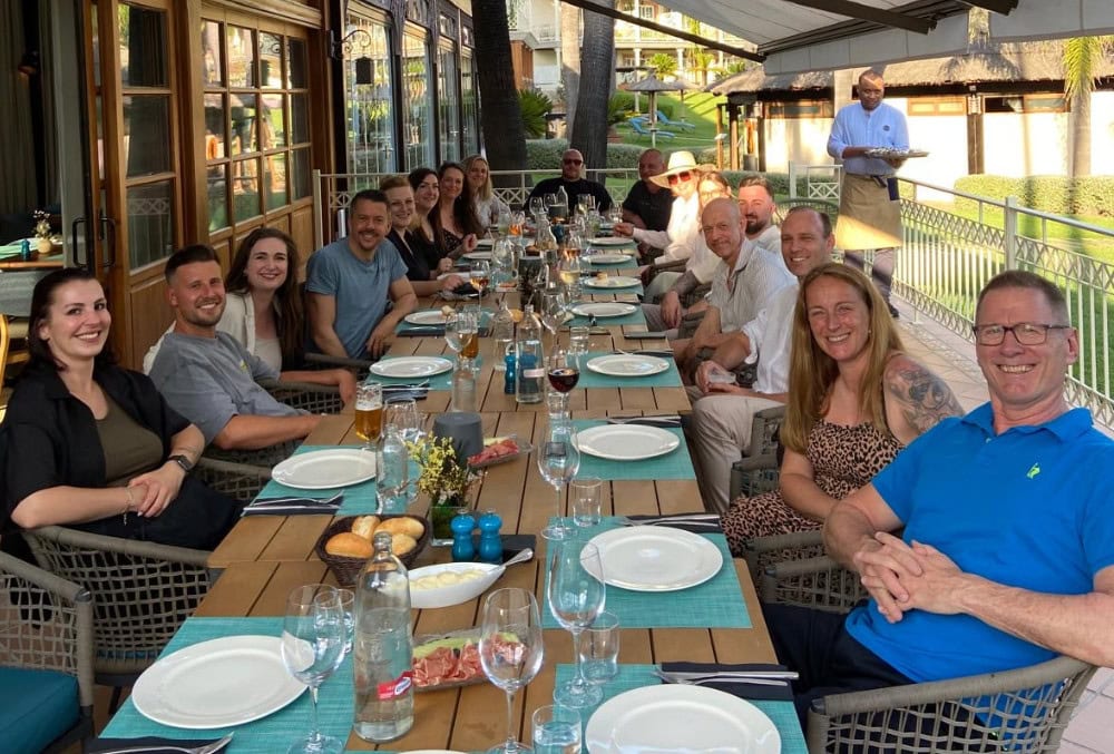 Gruppe von 17 Personen beim Geschäftsführertreffen 2025 auf Mallorca. Die Teilnehmenden sitzen lächelnd an einer langen, gedeckten Tafel auf der Terrasse eines mediterranen Hotels.